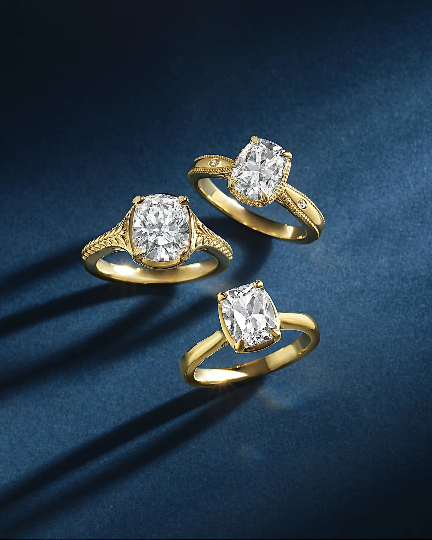 Three yellow gold engagement rings: an oval with hand-engraved shoulders, a cushion with milgrain and bezel-set accents, and a cushion solitaire